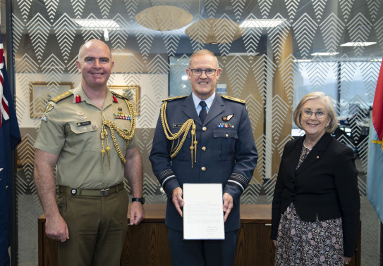 Three people stand in front of a glass wall with patterns on it. The New Zealand flag to the left. Chief of the New Zealand Defence Force Air Marshal Kevin Short (centre) was presented the commendation by Defence Adviser Colonel Neil Peake (left) and High