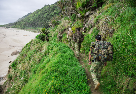 Soldiers trek in military gear with packs. A beach is to the left and hills stretch into the distance on the right.