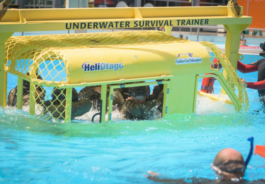 Soldiers in uniform and helmets are lowered into a pool by a crane, while sitting in a small metal helicopter simulator 
