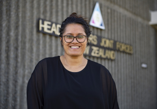 Miss Keri Brooking, 2022 Civilian of the Year, stands in front of the Headquarters Joint Forces New Zealand sign. She is wearing a black top and is smiling at the camera. 