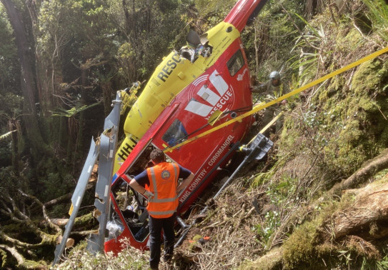 A red and yellow Westpac Rescue helicopter crashed in dense bush with a person inspecting the helicopter in the foreground. The area of bush around the helicopter has been cut and cleared, ready for recovery.