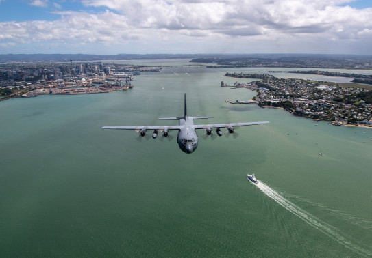 The grey C-130H Hercules aircraft flies over Auckland Harbour with the Auckland Harbour Bridge and SkyTower in the background.