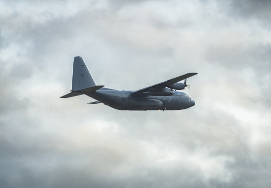 A rear-side view of the grey C-130 Hercules aircraft, with landing gear up shortly after taking off from Christchurch Airport. The clouds in the background are very textured and broken with a soft light glowing the edges.