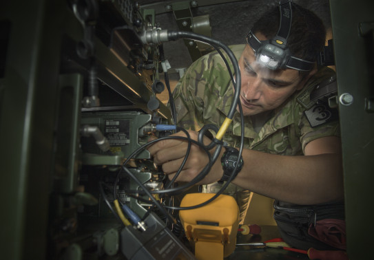 New Zealand Army's SGT Teaora Wright, an Electronics Technician working on electronics in a vehicle. He is wearing camouflage uniform and a head torch.