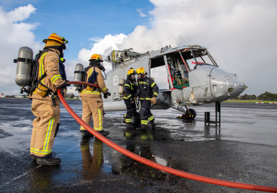 Four firefighters two in grey suits holding a red hose, and two in black suits, with yellow helmets tending to the shell of a Seasprite helicopter used in the exercise to simulate an aircraft crash.