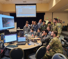 A number of uniformed military personnel stand in a room with lights on looking at various computer screens.