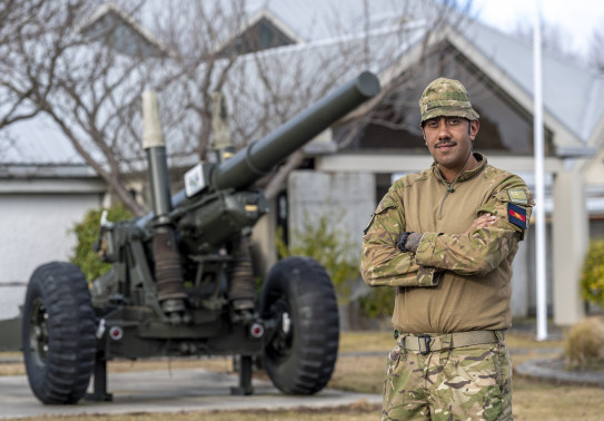 Staff Sergeant TJ Chapman, wearing fatigues standing cross armed in front of an L119, 105mm Light Gun, which is parked outside a headquarters building.