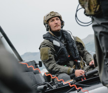 A soldier wearing full wet weather Army uniform including a life jacket and helmet is photographed between two other soldiers as they sit in small boats under grey skies.