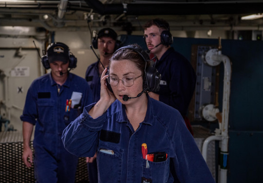 A woman wearing Navy blue overalls, glasses and a headset at work in the electrical part of a ship.