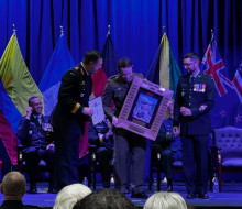 Major Prouse recieved a trophy on stage from two other military personnel. The lighting is very blue and flags line the back of the stage in front of a stage curtain.