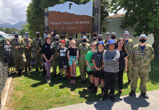 The officer cadets with some of the students and staff from Wairau Valley School. The uniformed personnel have masks on and it is a sunny day. 