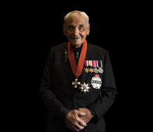 A man wearing a black suit and multiple medals stands in front of a black background.