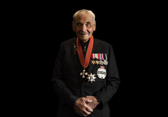 A man wearing a black suit and multiple medals stands in front of a black background.