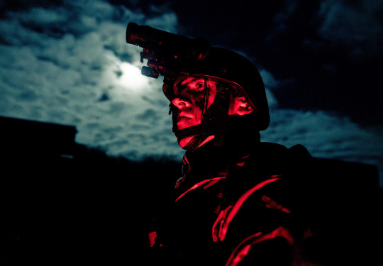 A soldier glows red while standing in front of moon-lit clouds wearing a helmet and Army uniform at night.