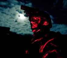 A soldier glows red while standing in front of moon-lit clouds wearing a helmet and Army uniform at night.