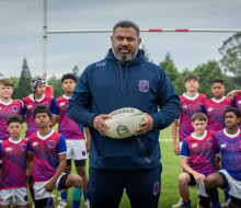 A man holds a rugby ball in front of a team of young players.