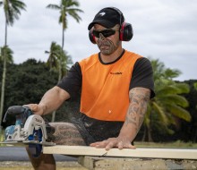 A soldier in high vis orange saws a piece of wood, The soldier is wearing a black peaked cap with an NZDF Kiwi logo on it, ear muffs and eye protection. It's a cloudy day. 