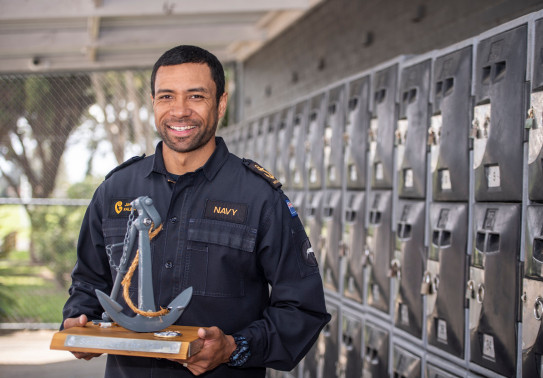 Man in Navy uniform smiles as he holds a trophy that resembles an anchor.