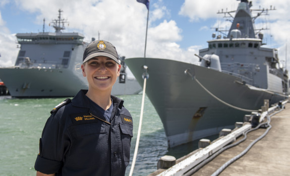 A sailor stands smiling on a sunny day, with some clouds. There are two ships in the background. 