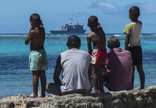 Fijian locals watch from the shore as a Seasprite helicopter lifts an under-slung load from the flight deck of HMNZS Canterbury. 