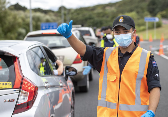 A navy sailor wearing high vis and a mask gives the thumbs up to a vehicle during a checkpoint inspection in Auckland