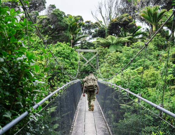Soldiers in a line, walk over an elevated swing bridge on the Heaphy Track. Trees reach upwards on either side.