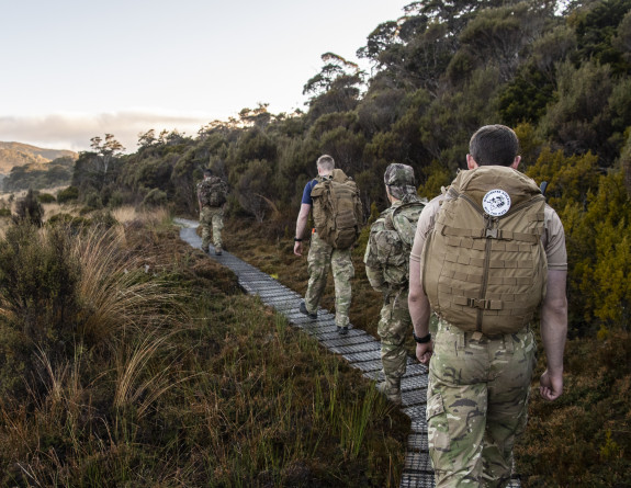 Soldiers walk in a line on a winding wooden boardwalk. The landscape to the left shows a wet tussock area and green hills stretching beyond.
