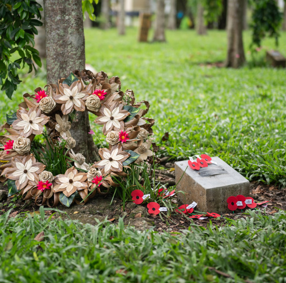 A wreath, poppies and a timber memorial plaque sit at the foot of a tree in the Henderson Field memorial gardens.
