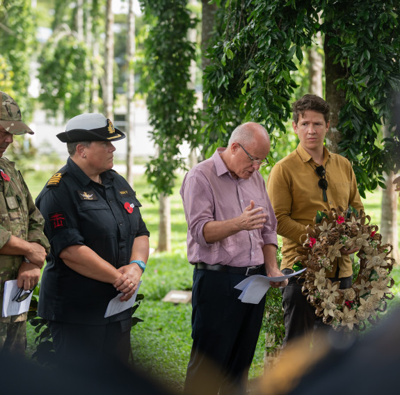 Between the shoulders of personnel, four people stand amongst the trees, two in NZDF uniform and two in civilian clothing. One man is reading from notes and another holds a wreath.
