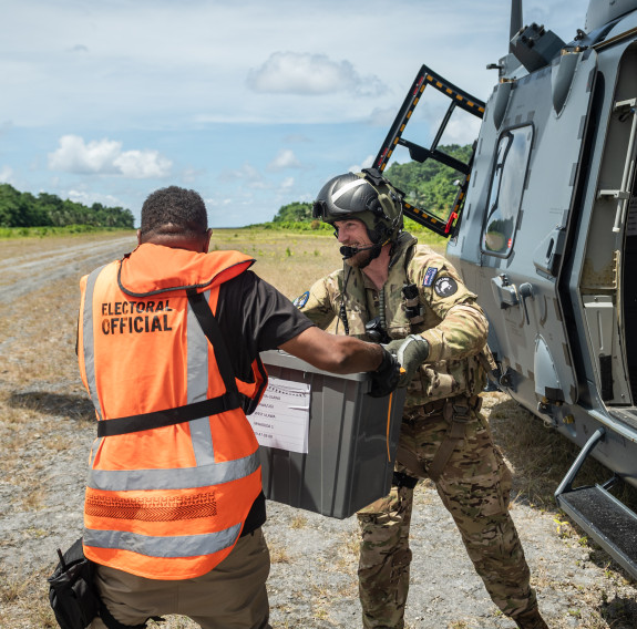 A helicopter loadmaster hands over a grey box to a man in a high visibility jacket with the words "Electoral official" on the back. On the right is the side of the NH90 helicopter and in the background a wide road, flanked by forrest travels to the horizo