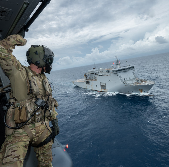 A loadmaster looks the side door of the NH90 helicopter to see the large grey HMNZS Canterbury cutting through the calm ocean. Clouds lap the horizon in the distance.