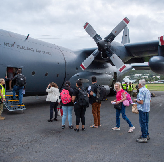People board the C-130H (NZ) from the front of the aircraft, the large wing and propellors above a queue of people. In the background personnel in high vis load cargo in the rear of the aircraft.