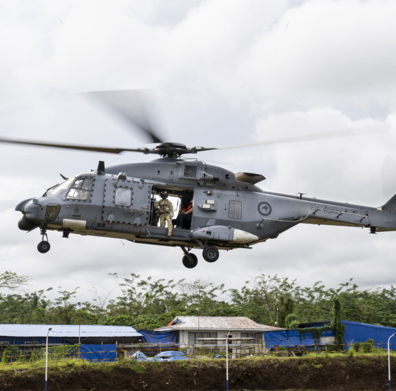 A grey helicopter after taking off flies just about tents and trees in the background.