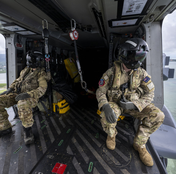 Two helicopter loadmasters sit inside a NH90 helicopter during flight. They are both looking out of the aircraft as they fly over the ocean with land in the background.