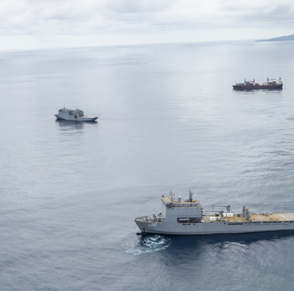 Two military ships and one cargo ship at sea, taken from the air. In the background the horizon meets land.