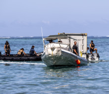 Two rigid hull inflatable boats manoeuver a shipping container at sea. The container has been floated using rigid pontoons.