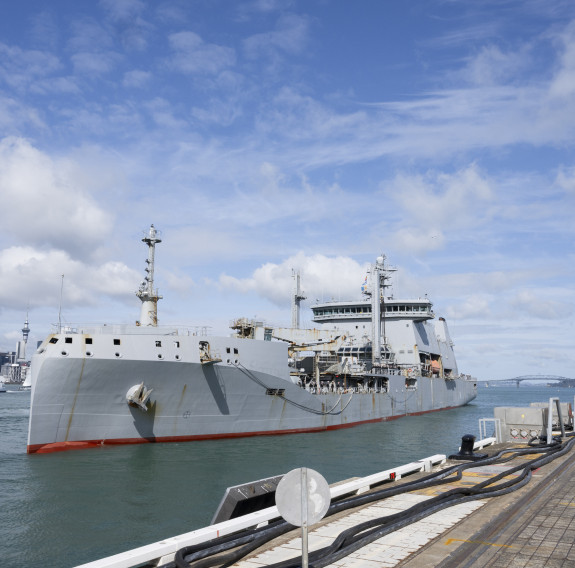 A large ship next to a wharf with a city skyline in the background. The sun is shinning and scattered cloud on blue sky is in the background.