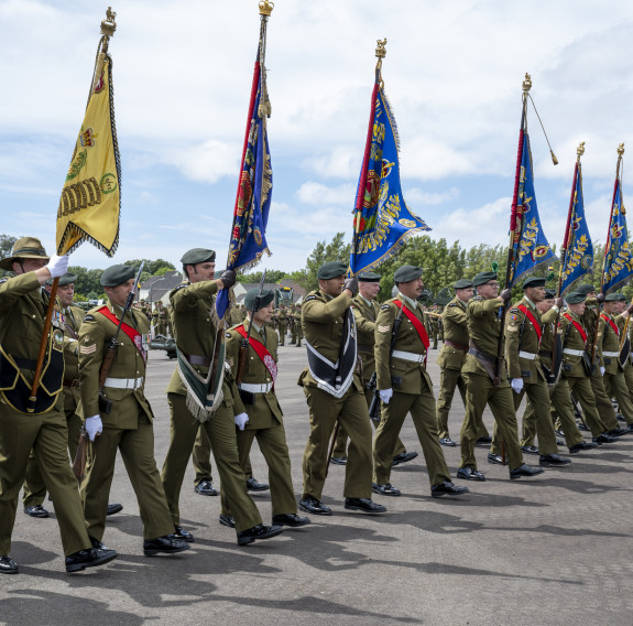 Soldiers in a line holding ceremonial flags (Colours) march forwards on a parade ground.