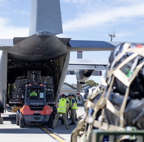 In the background to the left is the open rear end of a C-130H Hercules being loaded with a pallet by a forklift. In the foreground to the right is a pallet of aid secured by cargo webbing.