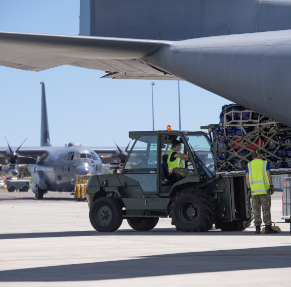 A military green forklift loads a pallet of aid into the rear of a C-130H Hercules. Another C-130H is visible in the background.