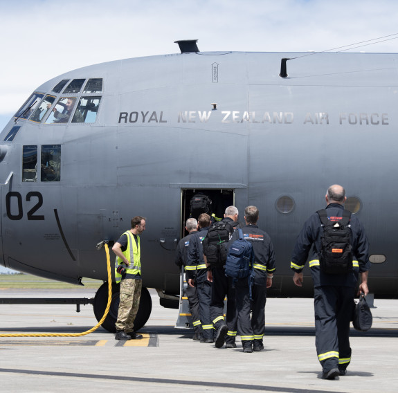 Urban Search and Rescue personnel board a C-130H Hercules through the forward door, watched by an RNZAF ground crew member wearing a fluorescent yellow high-vis vest.