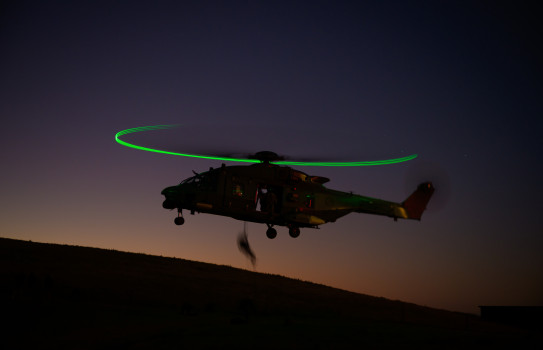 A helicopter with green lights on the rotors creates a halo as a police officer fast-ropes out towards a hill. In the background are the dim colours of sunset, silhouetting the helicopter and officer.