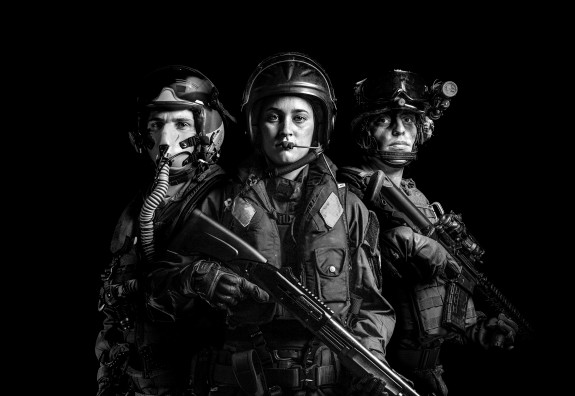A black and white photo of three NZDF personnel. The left is Air Force and is wearing a flying helmet and mask. In the middle the personnel is Navy and wearing a lifejacket and helmet with a headset. The person on the right is soldier and wearing headgear
