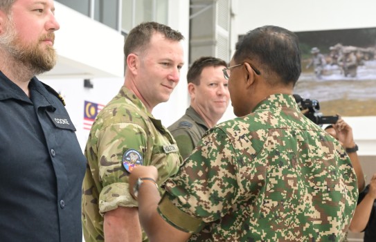 A man in Malaysian army camouflage attaches a round patch onto a velcro patch on the arm of a New Zealand solider, also in camouflage.
