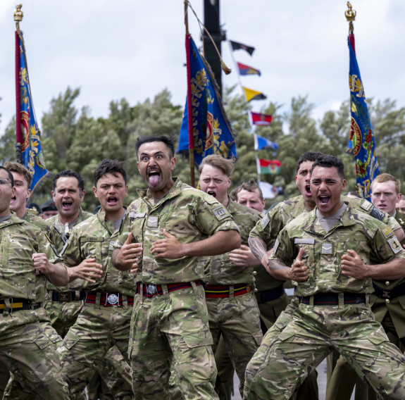 Soldiers perform a haka, ceremonial flags and trees appear in the background.