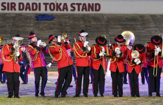 Army band members in red jackets and black trousers play musical instruments in front of an empty stand