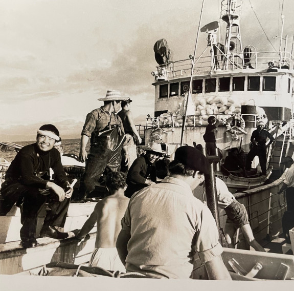 A sepia historic photo of a Japanese fishing vessel, one fisherman smiles at the camera as the others mill around.