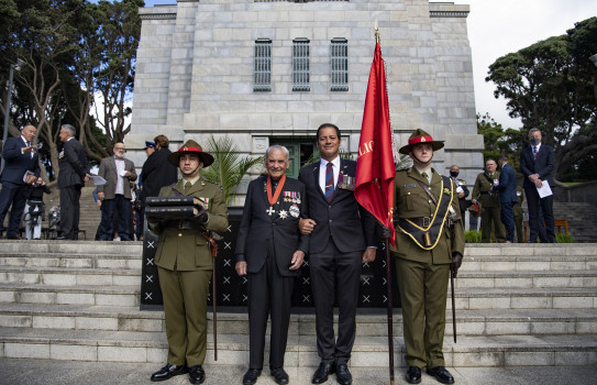 Four people stand in front of a war memorial. Two wearing suits and two wearing NZ Army uniform.