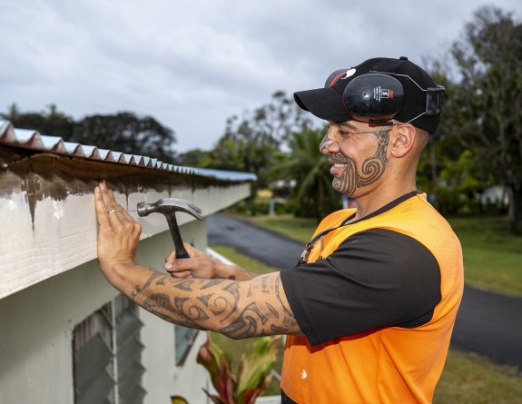 A solider in a orange high vis tshirt hammers a nail into a piece of wood. The image is taken side on and the soldier is smiling. 