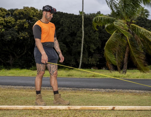 A soldier in a high visibility tshirt and shorts measures a piece of wood with a tape measure. 